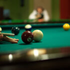 a photograph of a billiards table looking across the surface at table level. balls are visible all over the table. in the foreground, a hand and a pool cue line up for a shot next to the 7 and the cue ball. in the blurry background, another figure is resting their forearms on the edge of the table and looking at the person lining up the shot