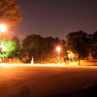 A color photograph of a deserted parking lot just after dusk. The lot is lit by two streetlights, which cast the entire picture into an orangy glow. The lot is framed by trees that have been turned black, orange, and green by the streetlights. A row of black trucks are facing out from the left side of the lot.