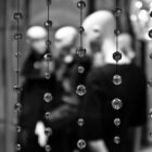 A black and white photograph of what looks to be a screen of beads, threaded onto string. In the background there are several white, bald, men that, all wearing black robes. The background is slightly blurred, while the bead-screen is in focus.