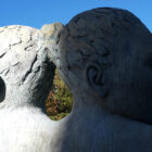 A color photograph of a statue of two, female, human heads, connected by their hair. The statue is made of gray stone. The right side of the statue is completely in shadow, the left side is fully in the light. The sky behind the statue is blue, with no clouds.