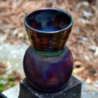 A color photograph of a multicolored pottery vase formed from a spherical form joined to a bowl form which rests on top of it. The vase is made with the traditional rakû style. It sits on top of a stack of two rectangular prisms made of concrete. The one directly beneath the vase is a dark slate grey, and the bottom concrete block is a light, chalky grey. The vase is framed by a blurred background of fallen leaves and brown pine needles with a small patch of green grass.