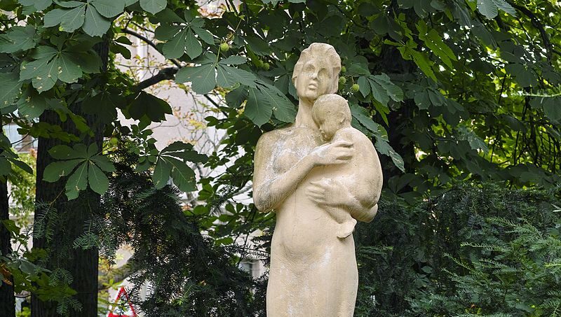 In color photograph of a statue in nature. It is a mother holding her child.