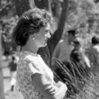 a black and white photograph of Jackie Kennedy in profile, with her arms crossed. There are people and trees in the background. She is wearing a short sleeved floral top and pearl earrings.