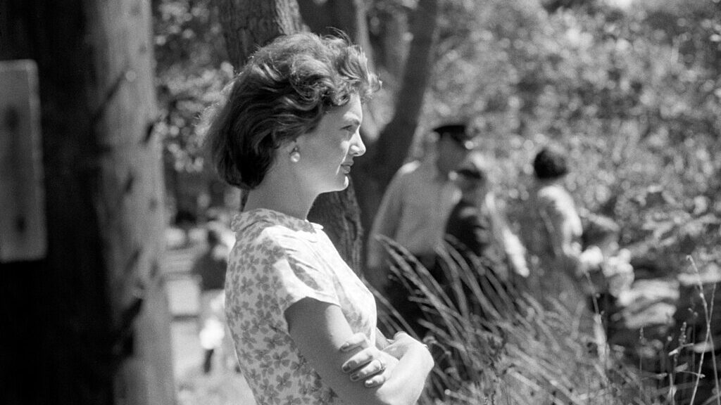 a black and white photograph of Jackie Kennedy in profile, with her arms crossed. There are people and trees in the background. She is wearing a short sleeved floral top and pearl earrings.