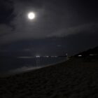 This is a dim photograph of a beach at night time with the moon peeking through the clouds.